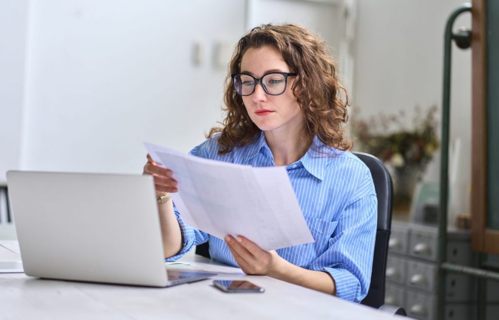 Girl with classes in blue dress shirt reading sheets of paper in front of computer