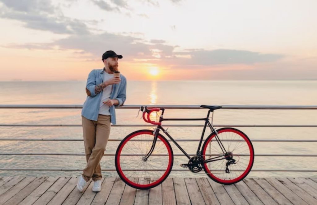 young bearded man with coffee in hand standing on a pier beside a bicycle with sunset in background