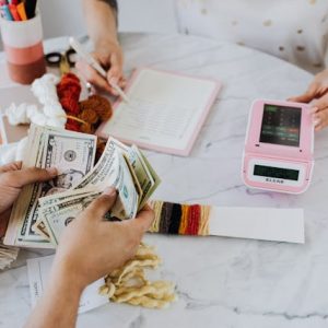 A person counting dollar bills while the other one is using a calculator and writing in a notebook