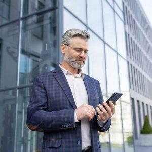 older man with gray hair in suit standing outside of a glass building looking at phone