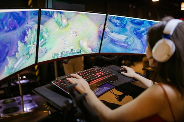 girl sitting at a desk with three computer monitors, keyborad, and mouse with headphones playing online games