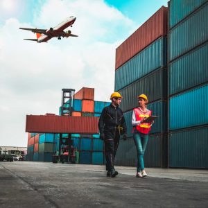 a plane flying over a ship with many containers stacked with 2 workers walking and talking