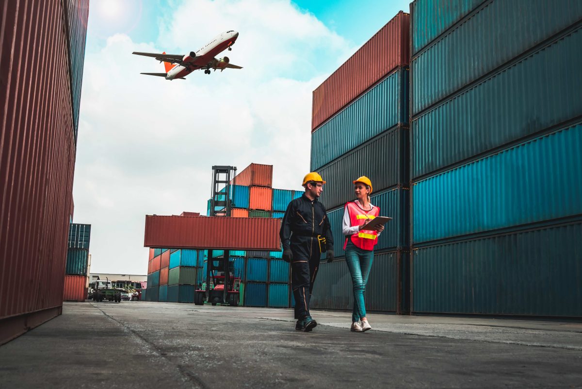 a plane flying over a ship with many containers stacked with 2 workers walking and talking