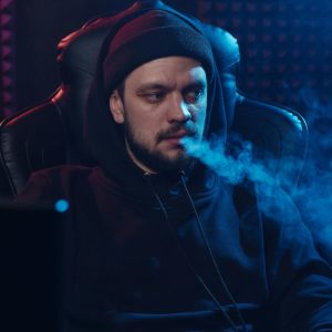 man in black hoodie sitting in a gaming chair with smoke coming out of his mouth