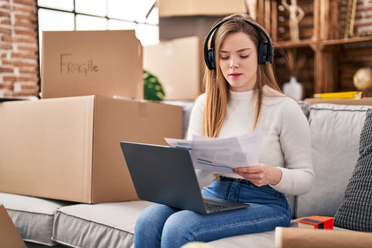 girl with headphones on, laptop in lap and moving boxes in the background.