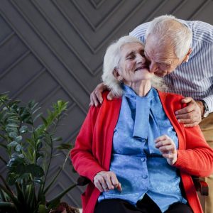 older woman sitting and older man giving the elderly woman a kiss