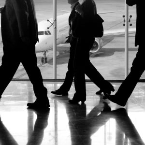 men in black pants and jacket walking in front of a wall of windows at an airport with plane in background