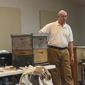 Stan Yeagley brought a bee hive to display as he talked about his work with bees. Items for beekeeping were also shown, including a frame spacer that maintains a consistent distance between frames within a hive box, a smoker to reduce the likelihood of bees becoming agitated, and a soft-bristled bee brush used to remove bees from a surface. He demonstrated the necessary apparel to wear in working with bees, including suit, gloves and head gear. Asked about often-seen white bee hives, he stated that the bees have shown no preference concerning the color of their hive homes.