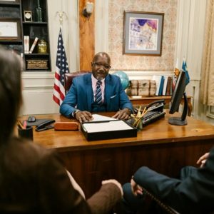 a person in a suit and tie sit behind a desk and talk to two other professionals