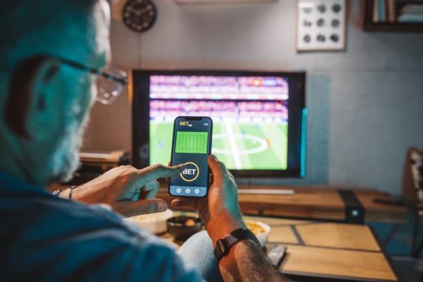 man holding a phone with sports betting and a soccer game on tv in the background