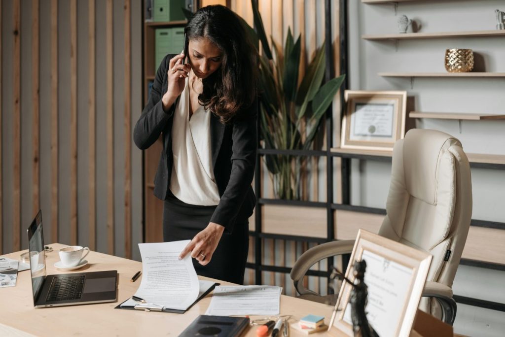 woman in black on cellphone and looking at papers on a desk