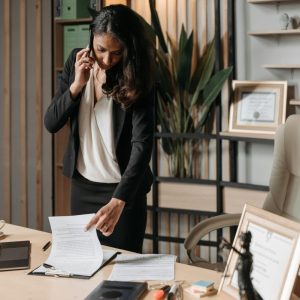 woman in black on cellphone and looking at papers on a desk