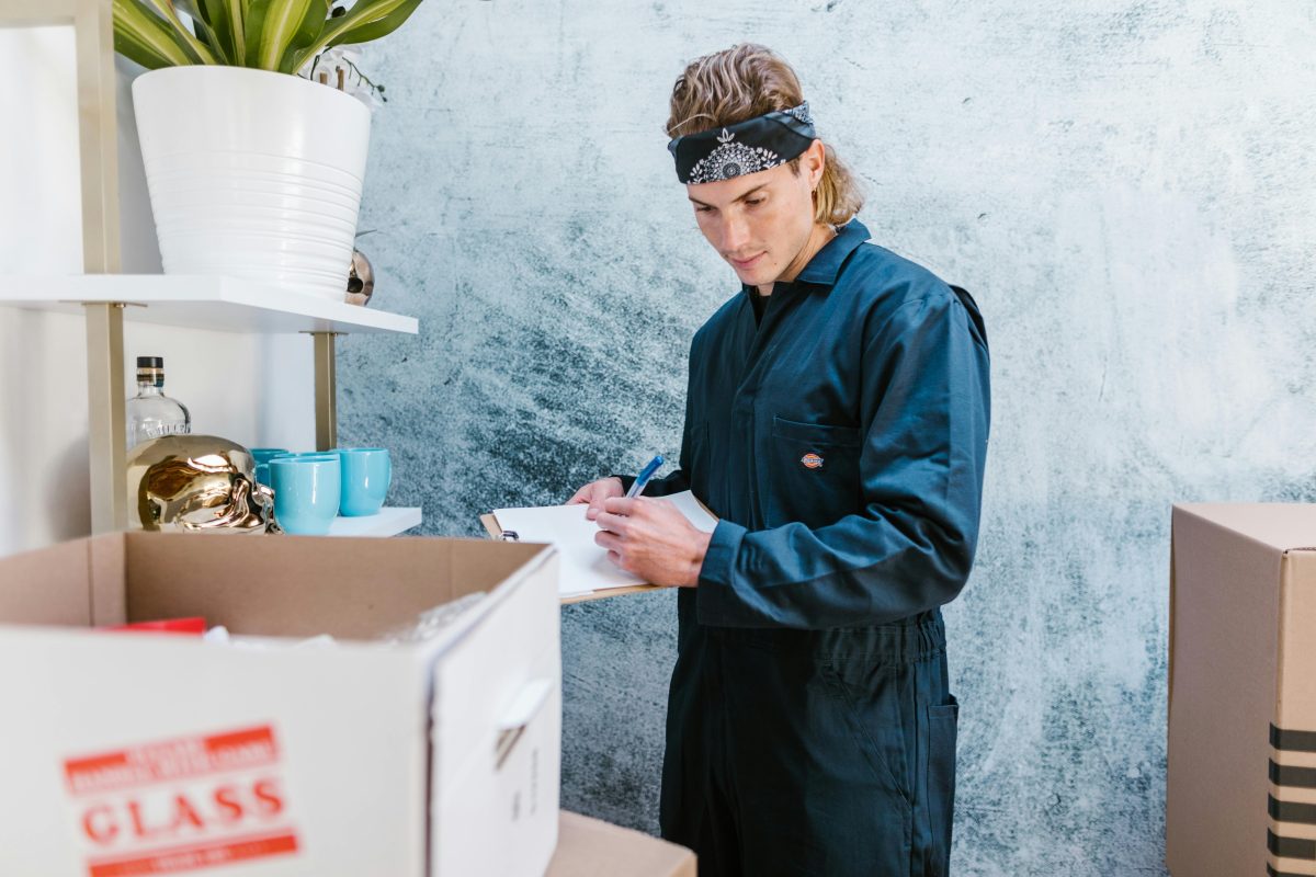 man in blue jumpsuit writing on a clipboard and looking at moving boxes