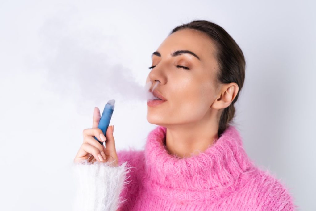A young cheerful girl in a bright colored sweater on a white background enjoys an electronic disposable cigarette with her eyes closed smokes releases smoke