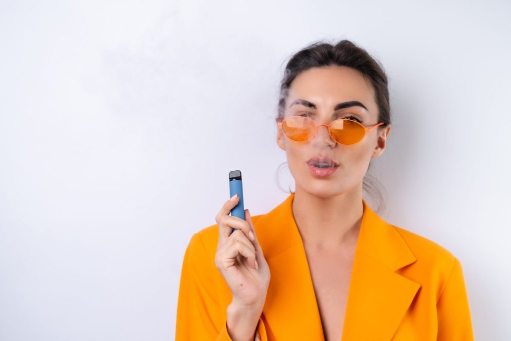 A young woman in trendy stylish glasses and a bright orange oversized jacket on a white background smokes an electronic cigarette