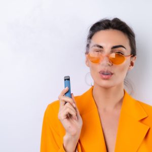 A young woman in trendy stylish glasses and a bright orange oversized jacket on a white background smokes an electronic cigarette