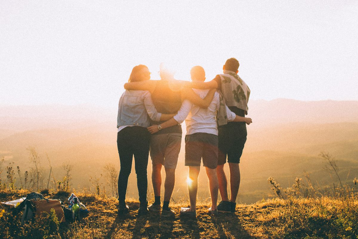 four people with arms around each other looking at sunset on mountain top