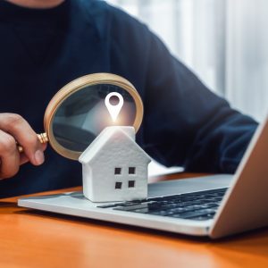 man holding a magnifying glass at a small house sitting on an open laptop