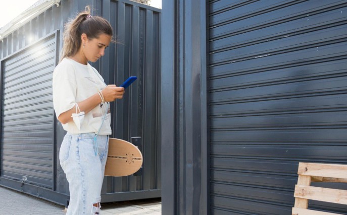 lady standing outside a storage unit holding a skateboard and a phone