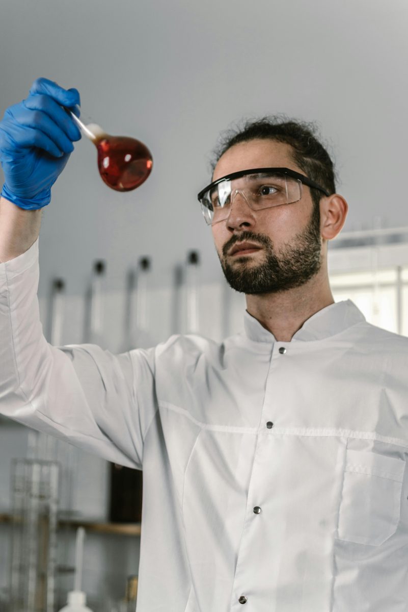 man in white lab coat and googles looking at flask with red liquid
