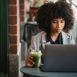 attentive-black-businesswoman-watching-laptop-with-beverage-in-cafe