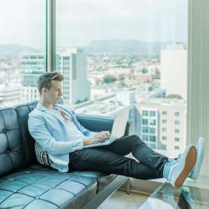 man sitting on couch looking at laptop with windows and city view in background