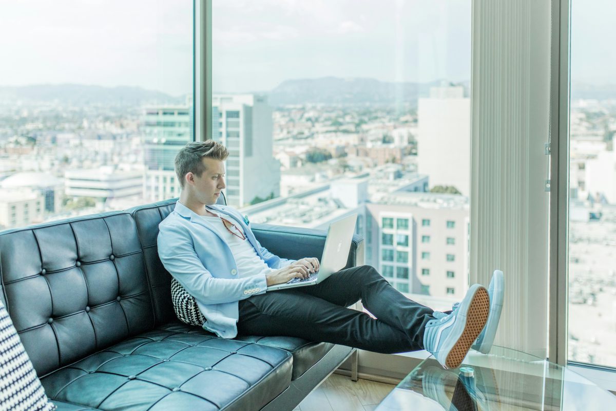 man sitting on couch looking at laptop with windows and city view in background