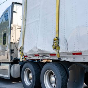 Beige industrial grade big rig semi truck tractor with broken dry van semi trailer fastened by sling standing on the city street waiting for possible repair or replacement of deformed trailer parts