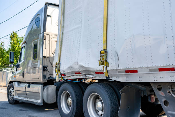 Beige industrial grade big rig semi truck tractor with broken dry van semi trailer fastened by sling standing on the city street waiting for possible repair or replacement of deformed trailer parts