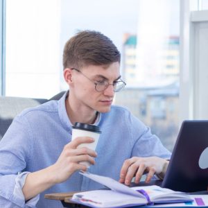 man with glasses at desk looking at laptop and holding a coffee