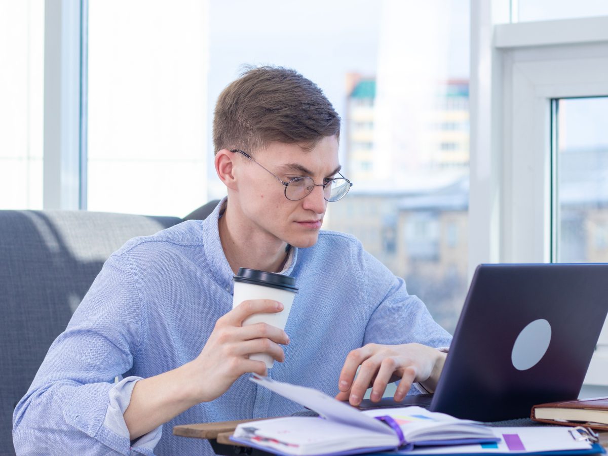 man with glasses at desk looking at laptop and holding a coffee