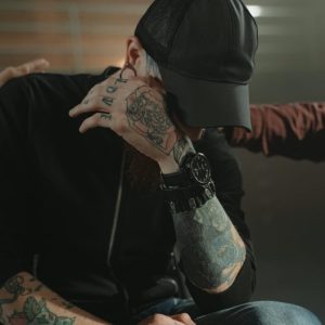 man with tatoos sitting in rehab covering eyes with hands patting him on the back
