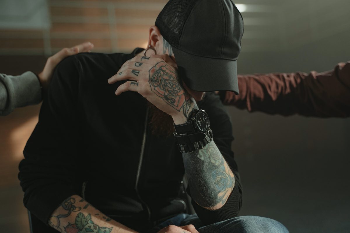 man with tatoos sitting in rehab covering eyes with hands patting him on the back