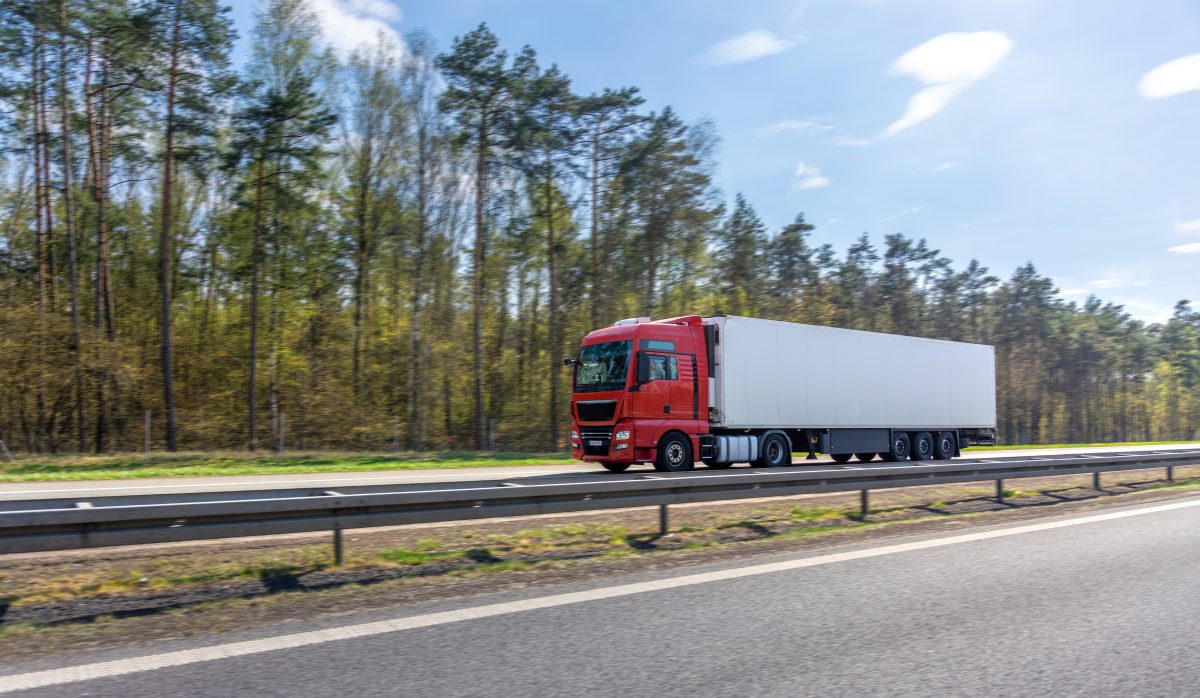 Red semi truck with white trailer driving on a highway commercial truck moves along the forest