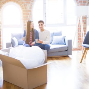 Young asian couple drinking a cup of coffee relaxing on the sofa at new home smiling happy around cardboard boxes from moving