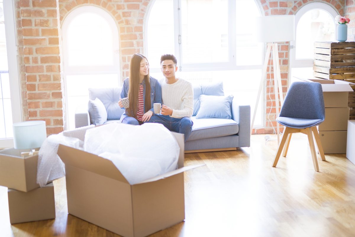 Young asian couple drinking a cup of coffee relaxing on the sofa at new home smiling happy around cardboard boxes from moving