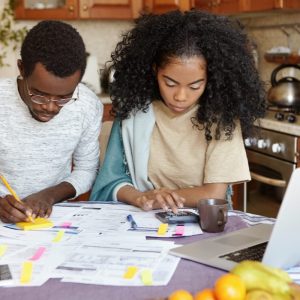 African man and woman sitting at kitchen table with papers and laptop pc, managing domestic finances together: wife counting on calculator while husband making notes with pencil. Family budget