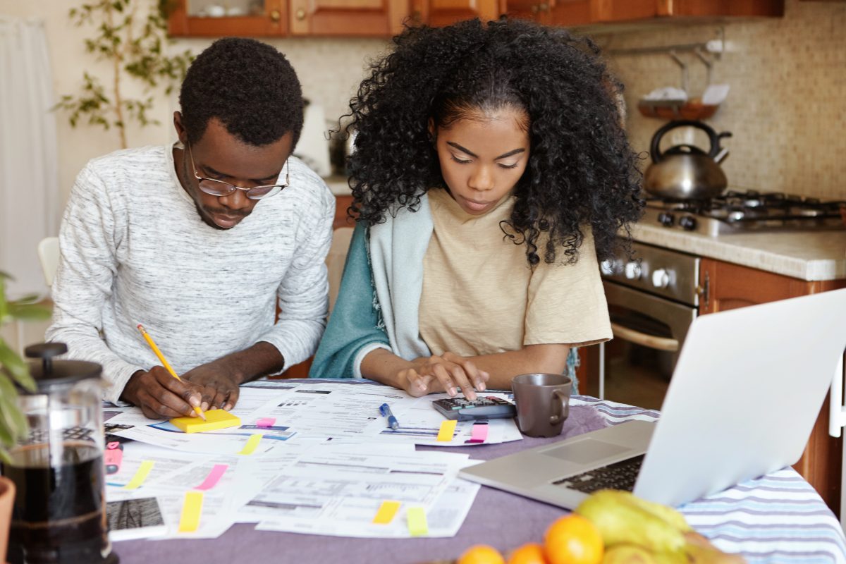 African man and woman sitting at kitchen table with papers and laptop pc, managing domestic finances together: wife counting on calculator while husband making notes with pencil. Family budget