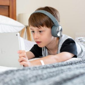 poy in blue shirt with headphones and tablet laying in bed
