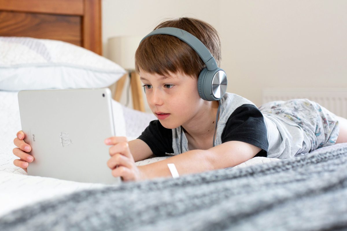 poy in blue shirt with headphones and tablet laying in bed