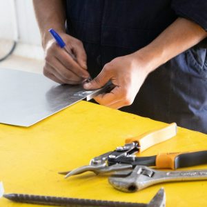 man holding a pen and writing on metal object with tools on yellow table
