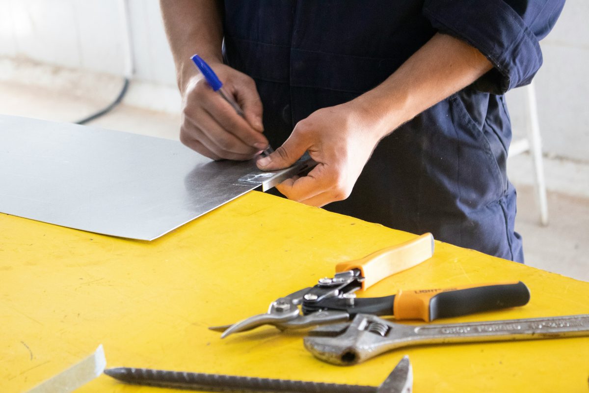 man holding a pen and writing on metal object with tools on yellow table