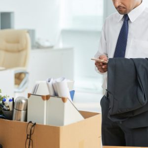 Unrecognizable man standing in office and using smartphone, with personal belongings in box