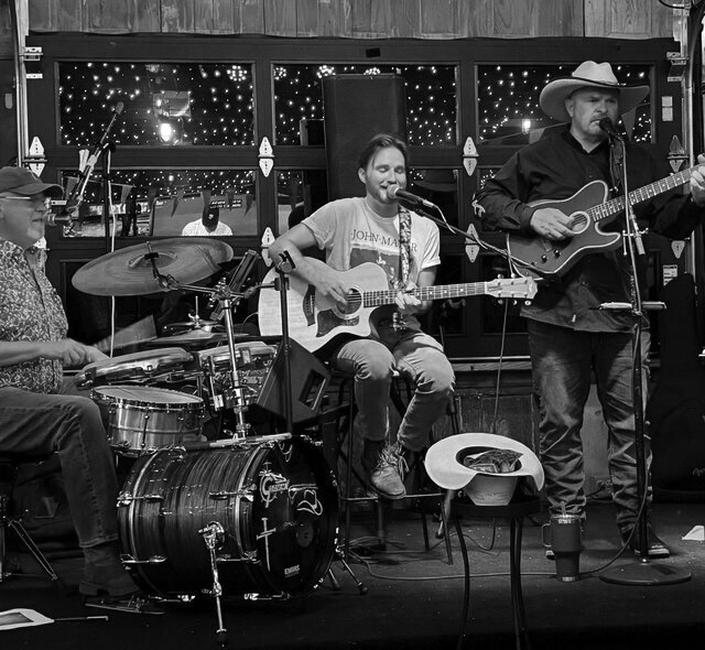 Black-and-white photo of three members of the band Cross Country performing on stage, with a drummer at left, a seated guitarist at center and a standing guitarist in a cowboy hat at right