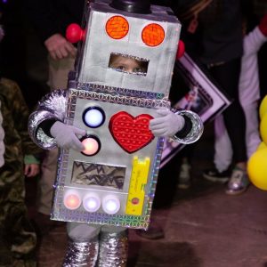 Child in a homemade robot costume with a red heart on the chest at a nighttime Halloween event