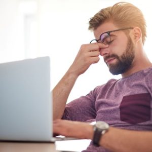 man rubbing eyes while sitting at a table looking at a laptop
