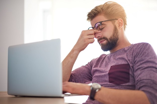 man rubbing eyes while sitting at a table looking at a laptop