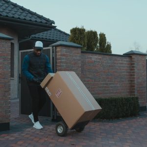 Professional delivery worker transporting package with hand truck in suburban setting.