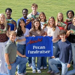 Attache’ Show Choir members posing on a lawn with a blue Attache’ Pecan Fundraiser banner