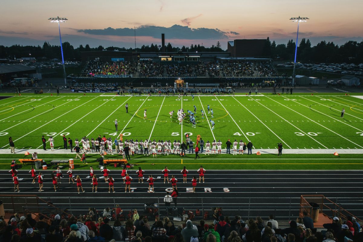 Players and a marching band on a lit high school football field at dusk, with fans in the stands
